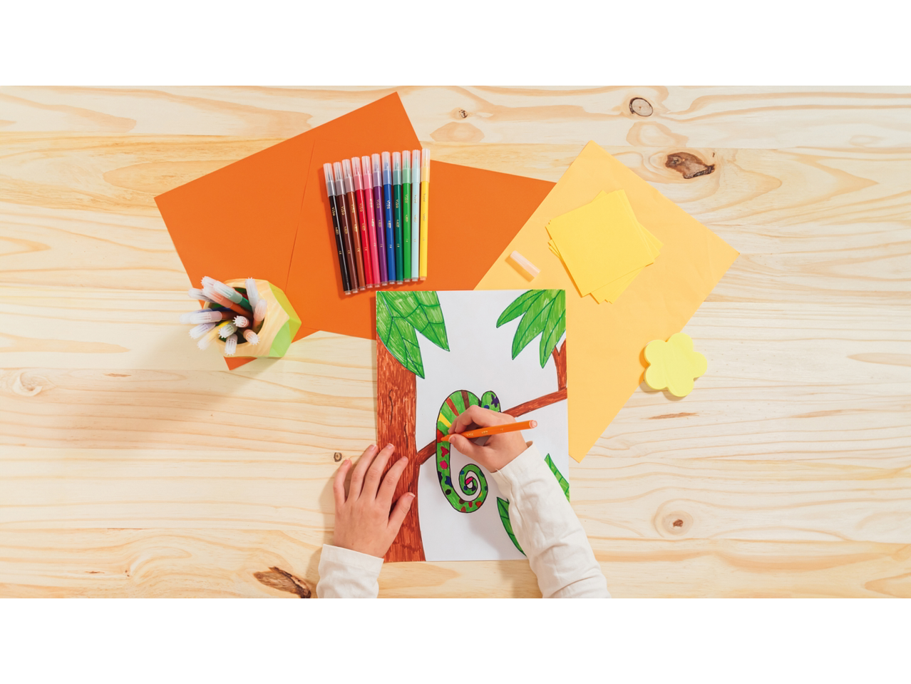 Child coloring a chameleon picture with markers, surrounded by stationery on a wooden table.