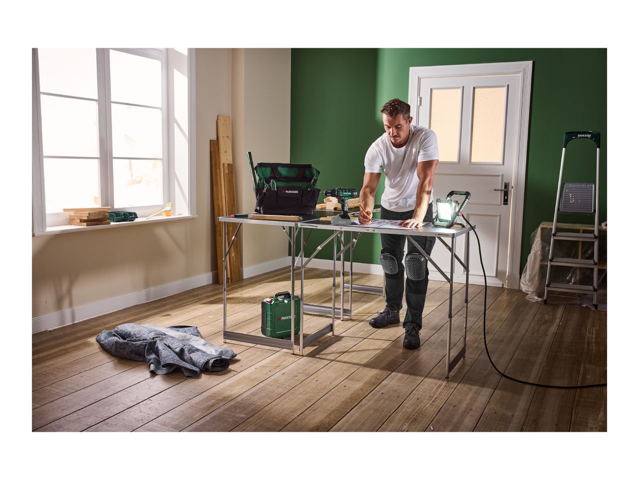 Man working at a folding workbench with a tool bag, drill, and work light.