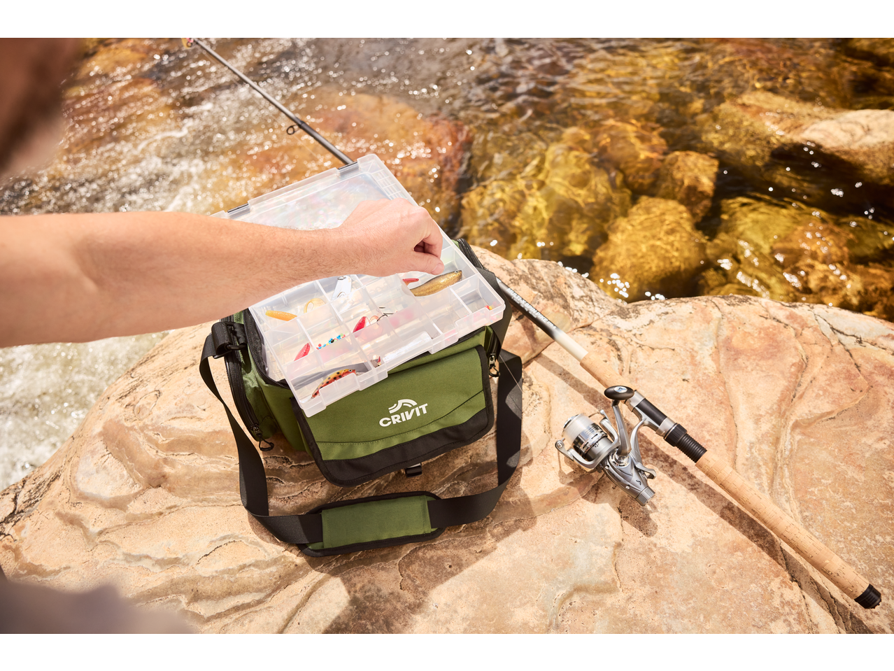 Echipament de pescuit CRIVIT: undiță, mulinetă și cutie cu momeli lângă râu.