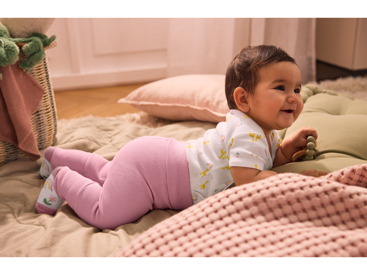Happy baby in a white bodysuit with giraffes and pink ribbed leggings, playing on a soft mat.