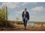 Man in een marineblauwe trenchcoat en donkere broek wandelt buiten bij een meer.
