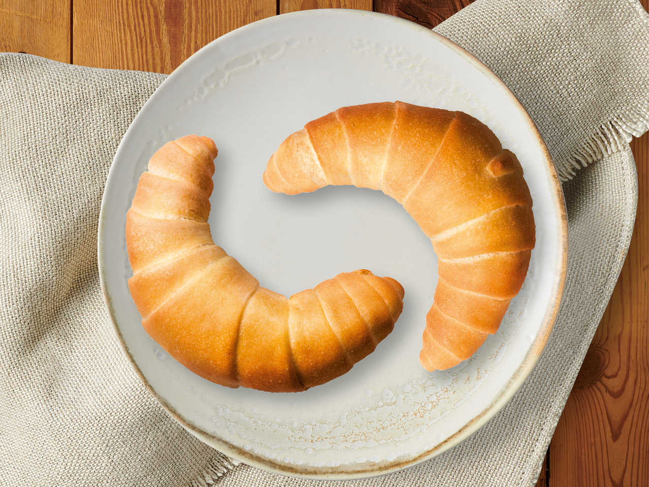 Два кроасана върху бяла чиния на дървена маса с ленена салфетка