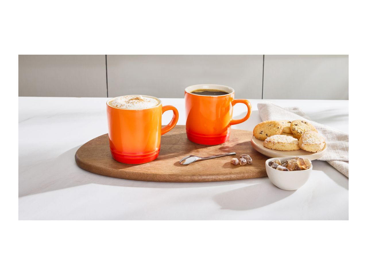 Two orange mugs of coffee, one with foam, on a wooden board with cookies and sugar.