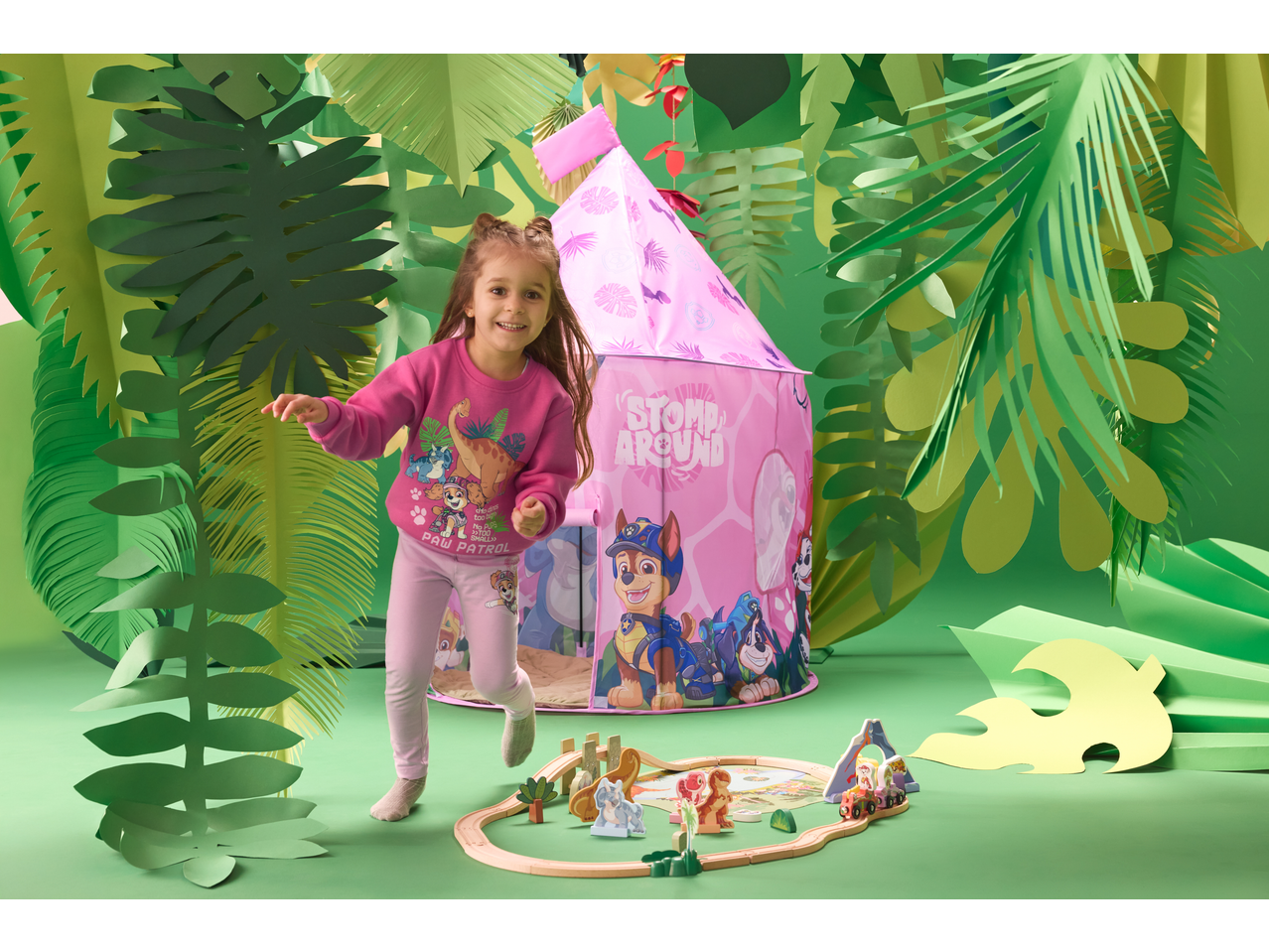 Menina sorridente em pijama de dinossauro e Patrulha Pata, a brincar junto a uma tenda de brincar rosa e uma pista de comboio de madeira.