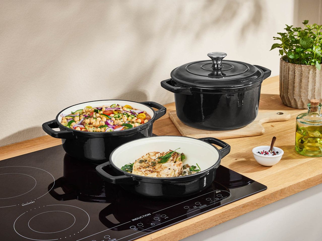 Black cast iron casserole dish set on an induction cooktop with food, next to a plant and olive oil.