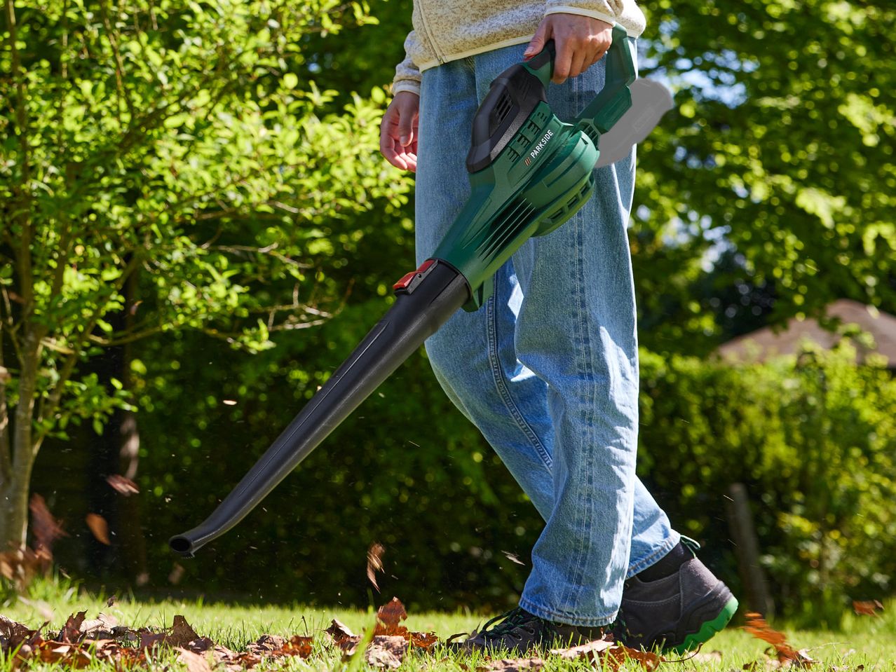 A person using a green and black leaf blower to clear leaves from a lawn.