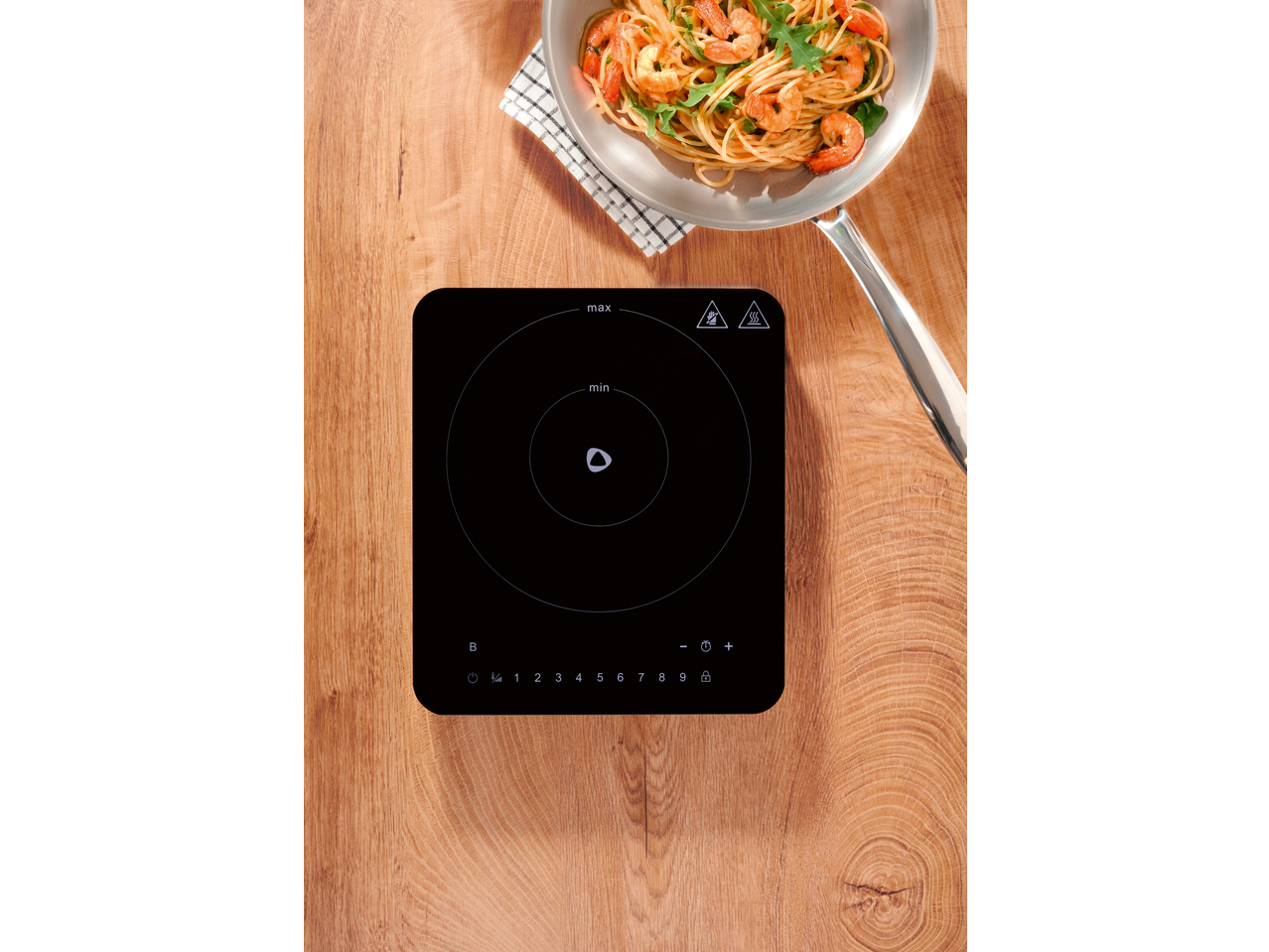 Placa de indução com uma panela de camarão e massa numa mesa de madeira.