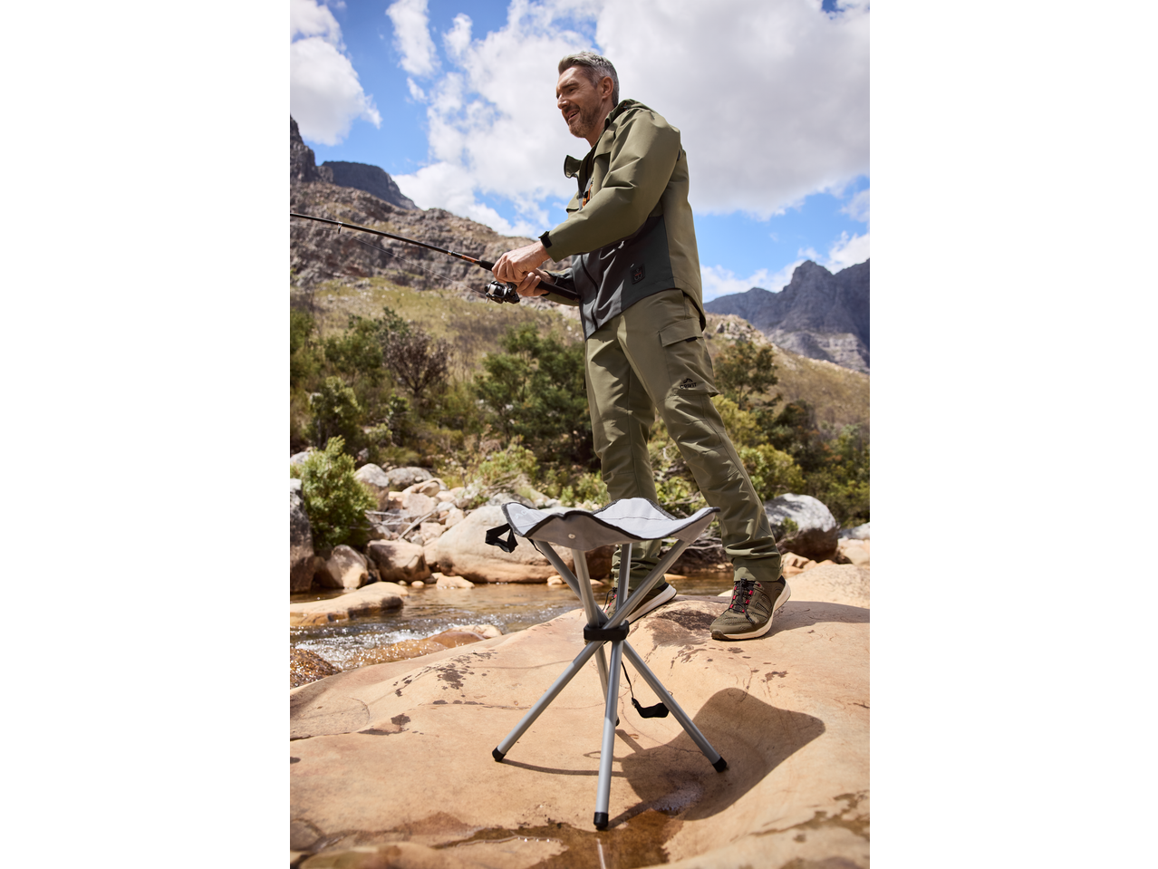 Muškarac peca sa CRIVIT setom, nosi ribolovačke pantalone i jaknu, pored sklopive stolice.
