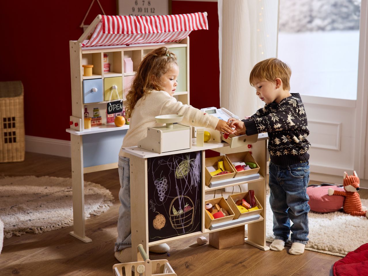Two children playing with a wooden toy market stall, featuring a scale and various play food items.