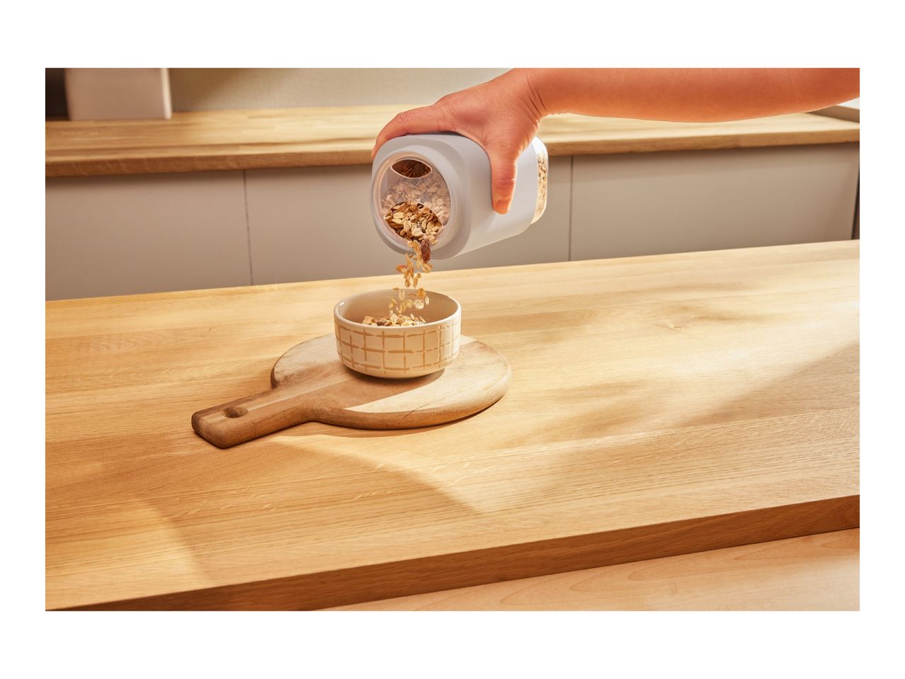 Hand pouring cereal from a dispenser into a bowl on a wooden cutting board.