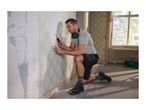 Man removing wallpaper with a scraper, wearing safety headphones, t-shirt, shorts, and work boots.