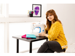 Mulher sorridente com uma máquina de costura E-ELEMENT BY PFAFF M3205 e tecidos coloridos.