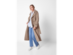 Een vrouw draagt een beige trenchcoat, een lichtblauw gestreept overhemd, jeans en witte sneakers.