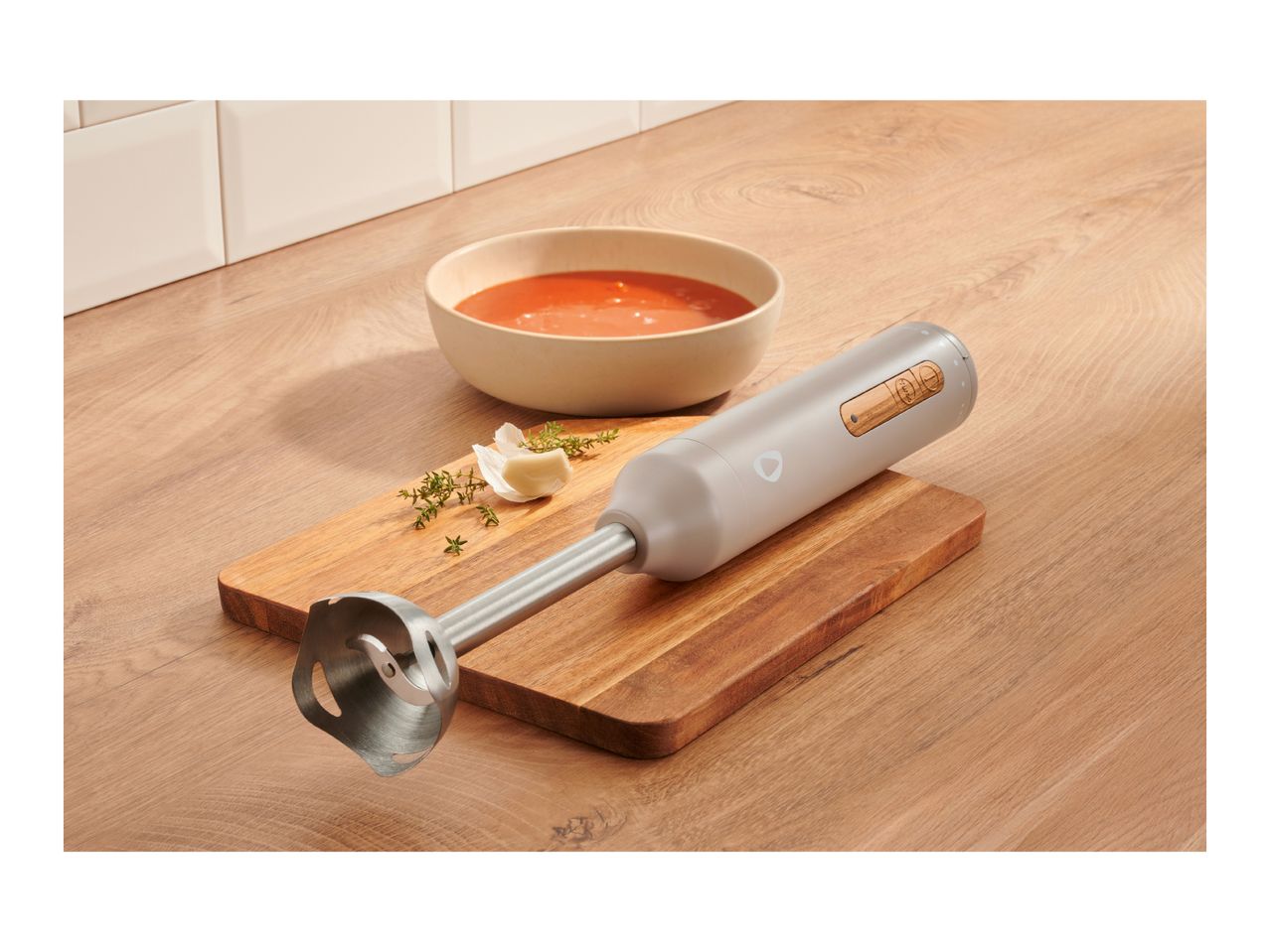 Immersion blender on a wooden cutting board next to a bowl of tomato soup, garlic, and thyme.