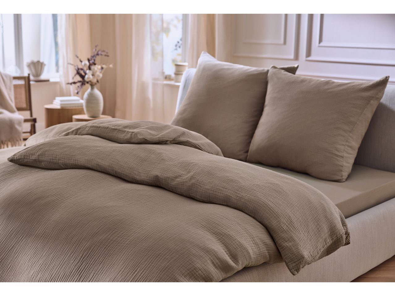 A bed with a brown duvet cover and two pillows, with a vase of dried flowers nearby.