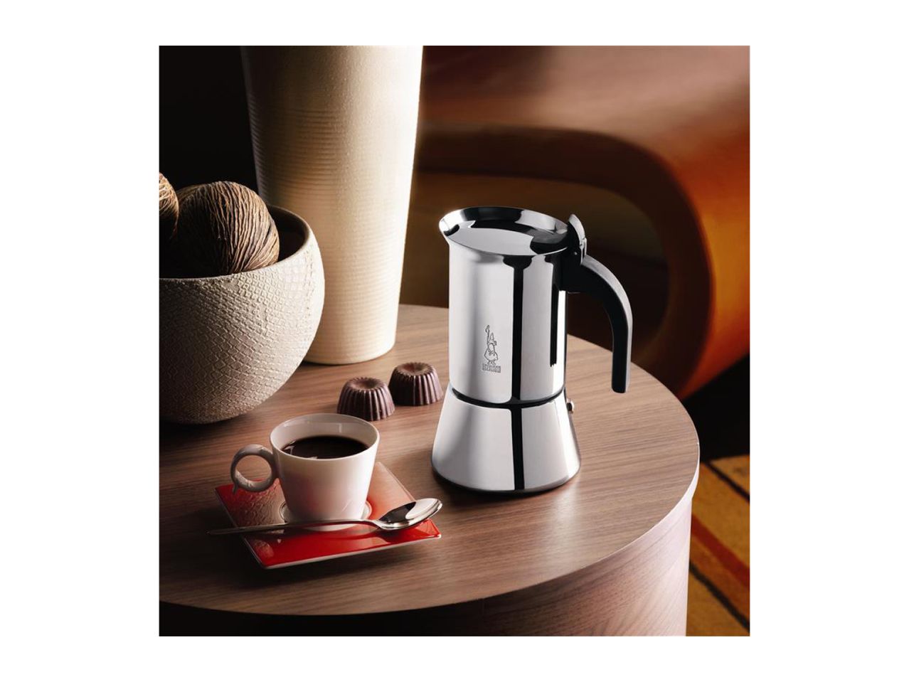 A stainless steel moka pot, a cup of coffee, and chocolates on a wooden table.
