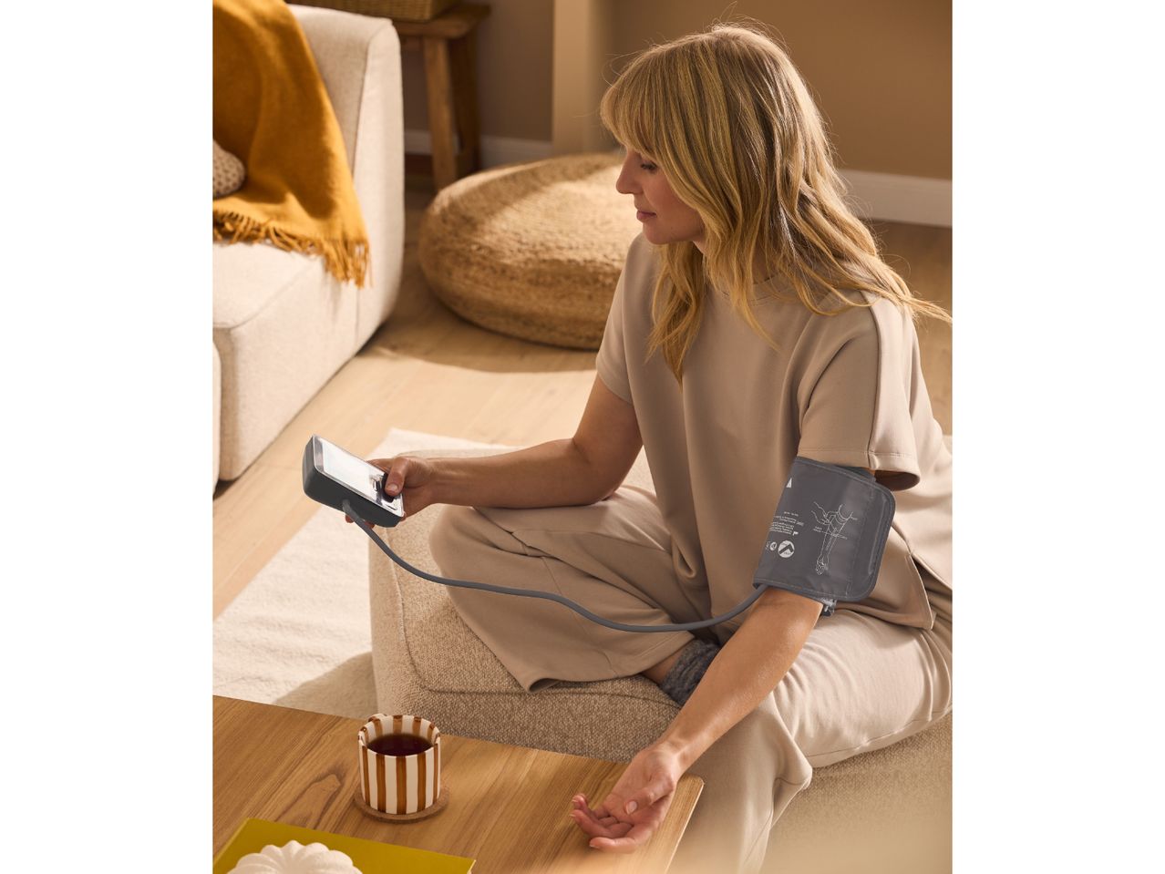 Woman measuring blood pressure at home, with a cup of tea on a wooden table.