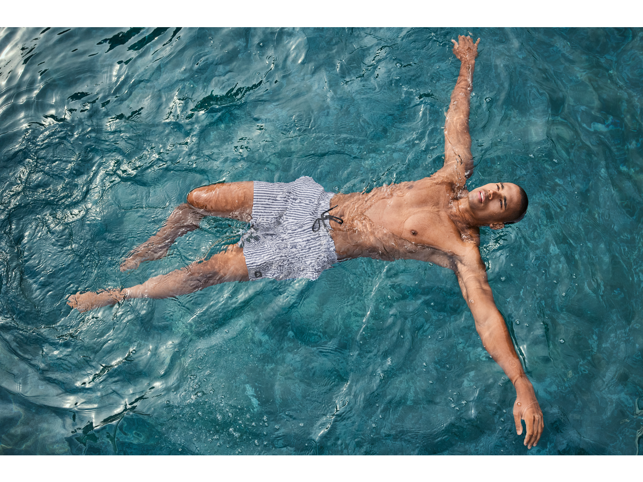 Man in striped swim shorts floating on his back in clear blue water.