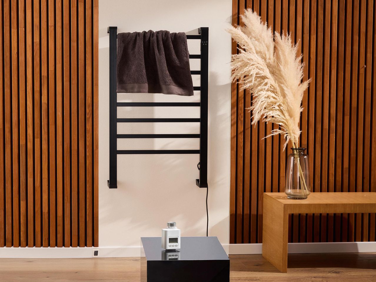 Fekete törölközőszárító radiátor barna törölközővel, fa panelekkel és pampafűvel.