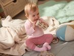 Happy baby in pink clothes playing with a wooden rattle on a soft blanket.