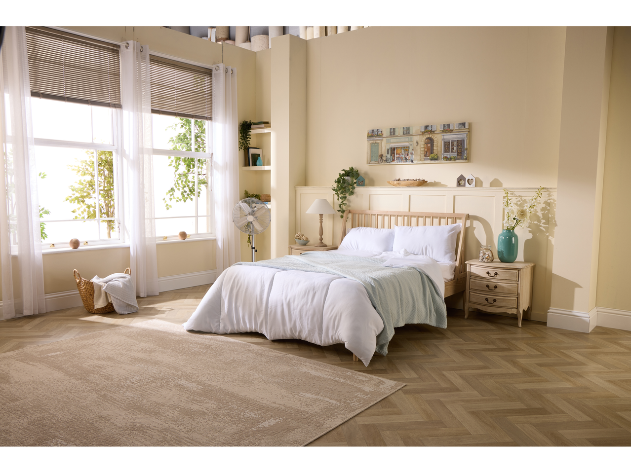A light and airy bedroom features a wooden bed with white bedding, a beige rug, and a standing fan.
