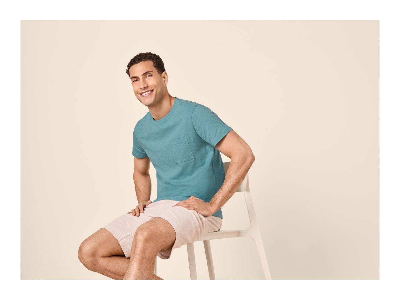 Man in a teal t-shirt and light shorts, sitting on a chair and smiling.