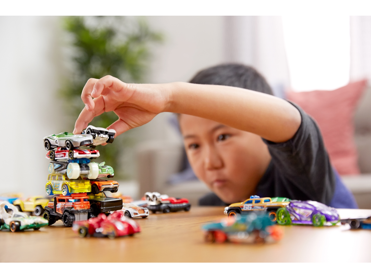 Child stacking Hot Wheels toy cars, with more cars scattered on a wooden table.