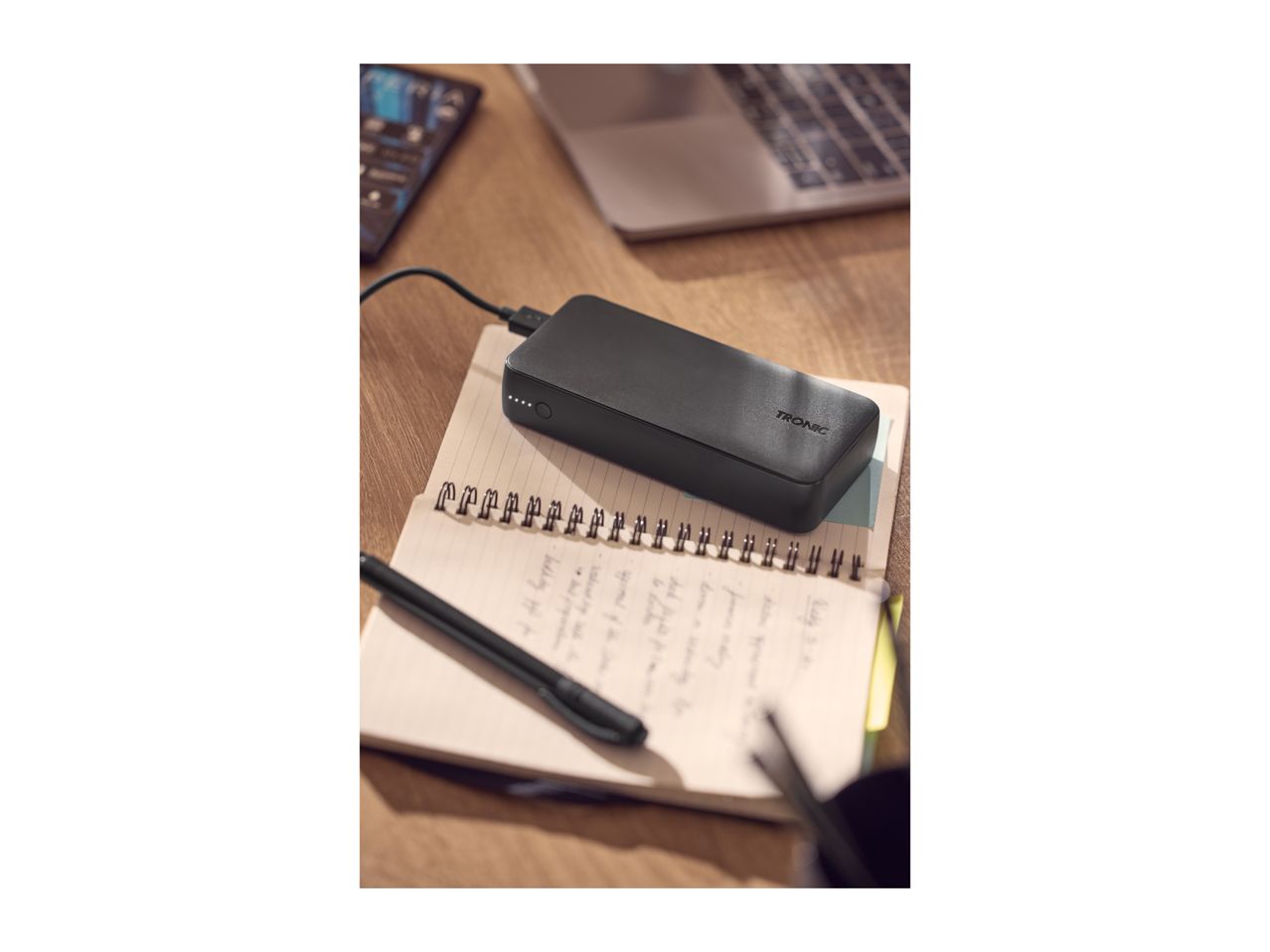 Black power bank charging on a notebook with a pen, laptop, and phone on a wooden desk.