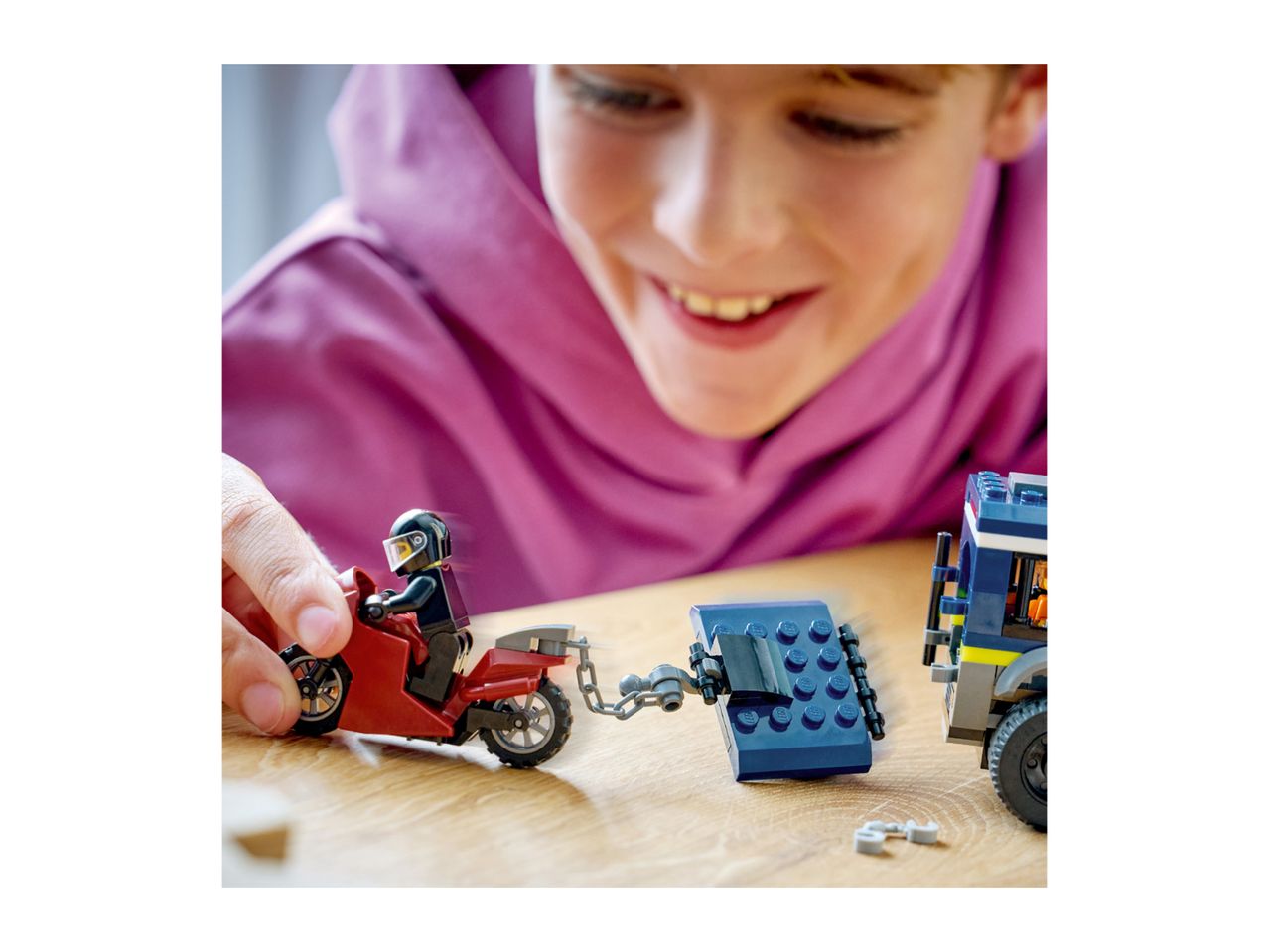 Smiling child playing with toy motorcycle and police truck on a wooden table.