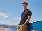Man in a navy polo shirt and khaki trousers, leaning on a boat by the sea.