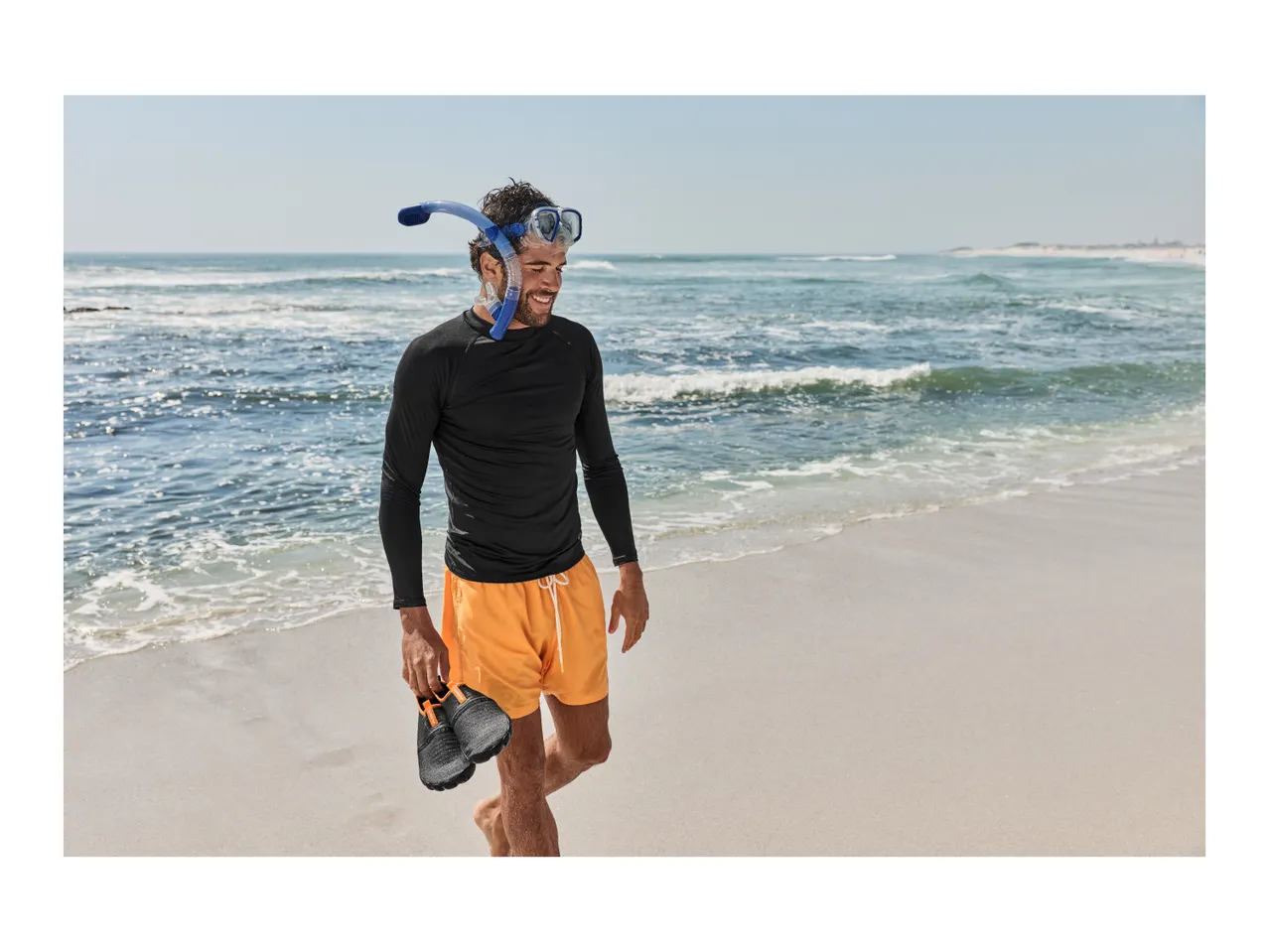 Man in black long-sleeve swim shirt, orange swim shorts, and snorkel mask walking on beach.