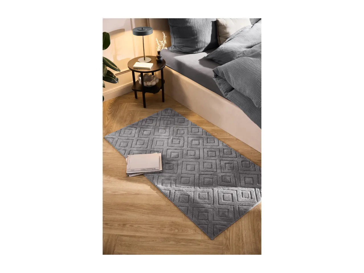 Grey geometric rug on a wooden floor next to a bed with grey bedding and a bedside table.