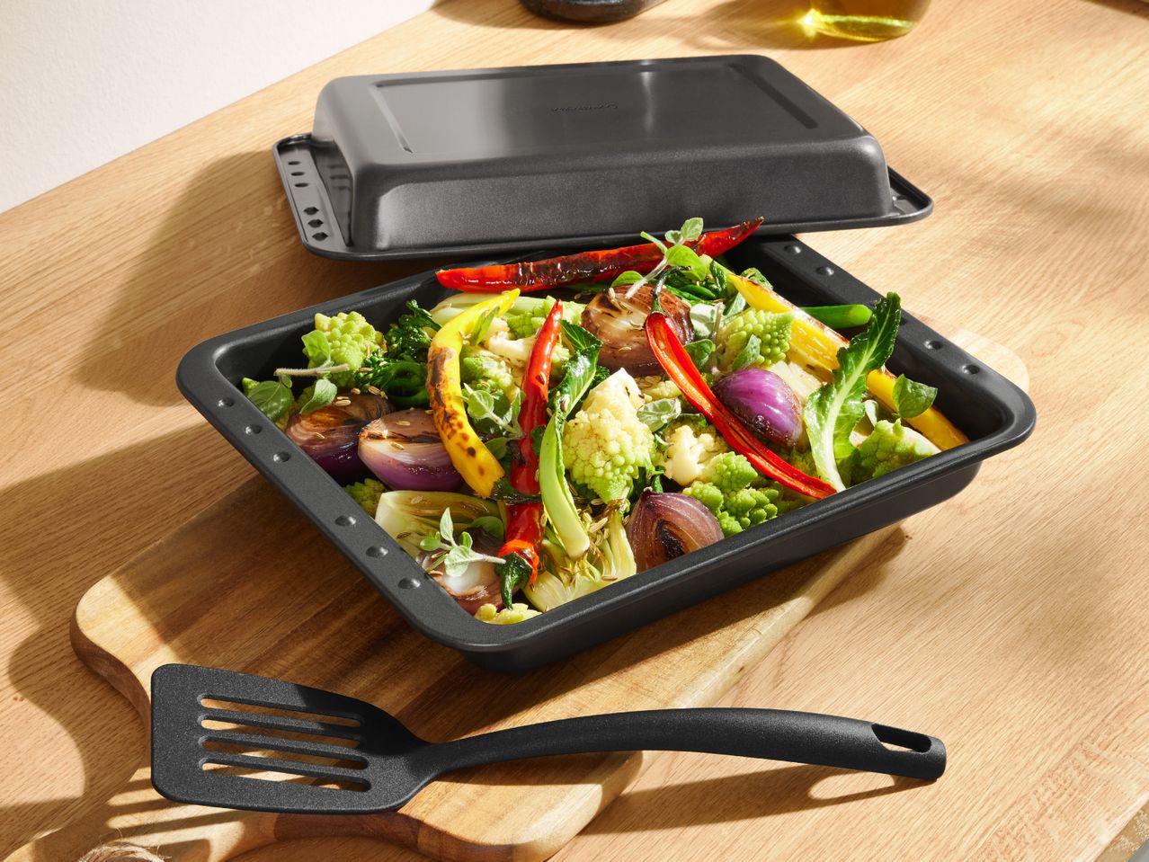 Roasted vegetables in a black baking dish with a matching lid and spatula on a wooden board.