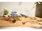 Tigelas de vidro com tampas cinzentas cheias de salada, sopa de tomate e azeitonas numa mesa de madeira.