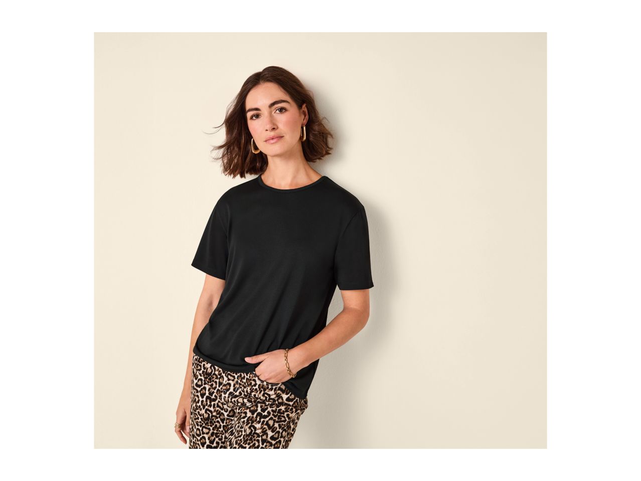 Woman in a black t-shirt and leopard print trousers, leaning against a wall.