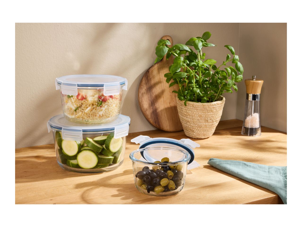 Glass food storage containers with food, basil plant, and salt grinder on a wooden counter.