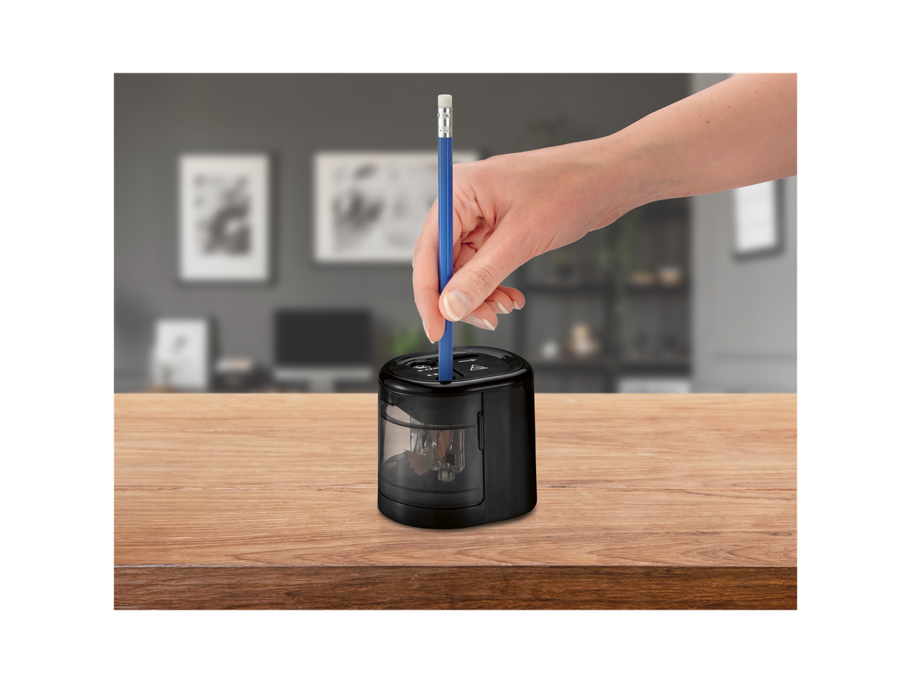 A hand inserts a blue pencil into a black electric pencil sharpener on a wooden table.