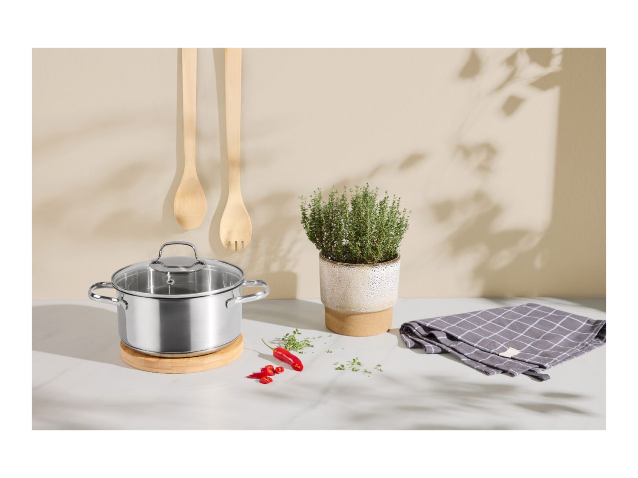 Stainless steel pot with lid, wooden utensils, potted herb, and checkered towel on a counter.