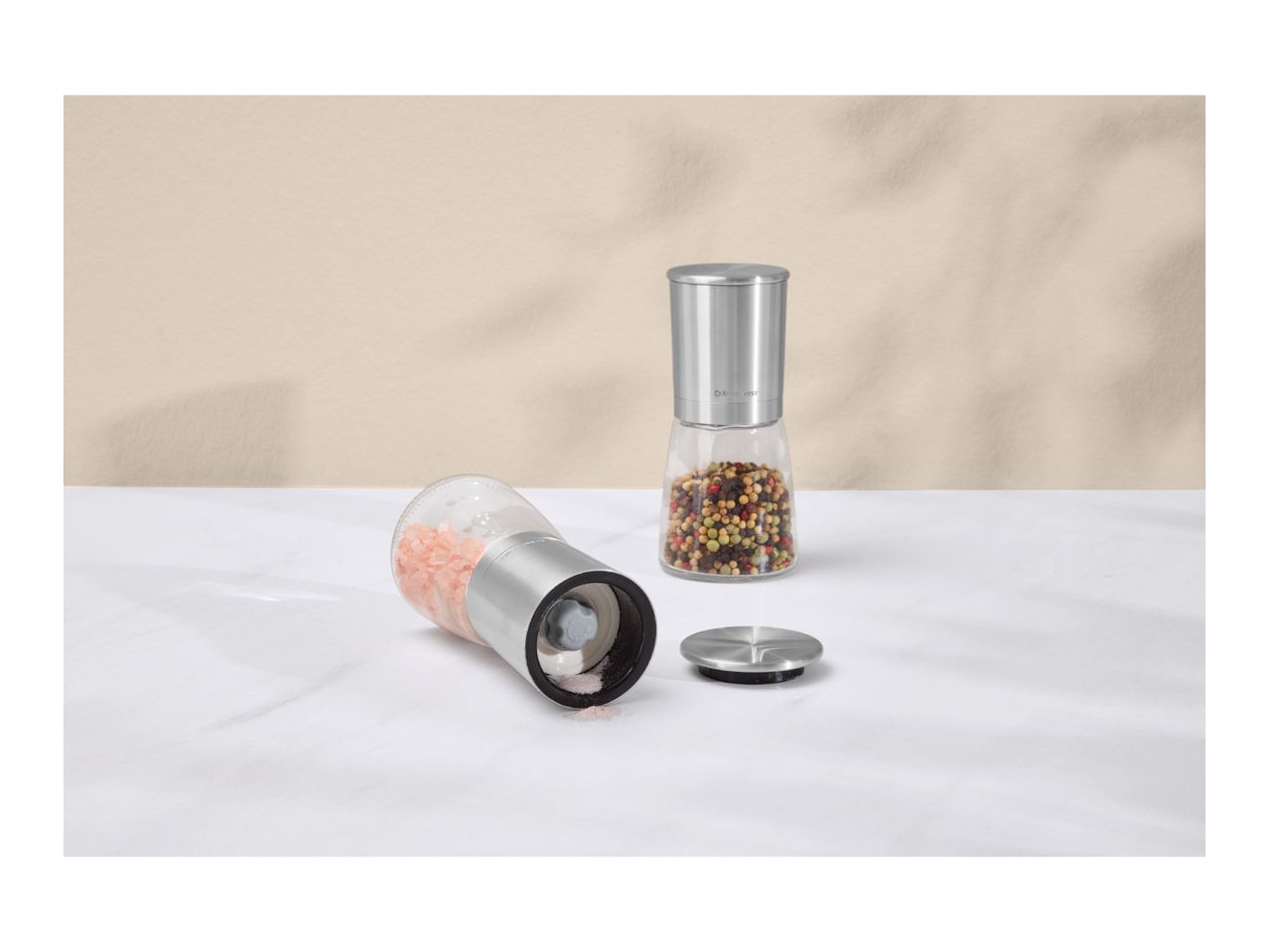 Two spice grinders, one with pink salt and one with mixed peppercorns, on a white surface.