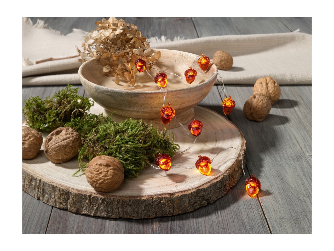 Acorn string lights, walnuts, and moss on a wooden slice, with a bowl and fabric.