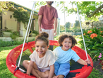 Two happy children in a red swing, with an adult standing behind them in a garden.