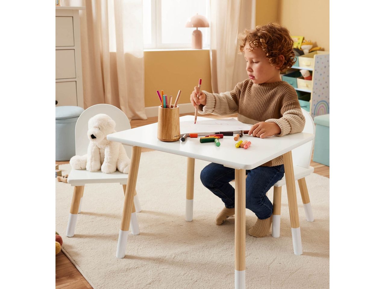 A young boy drawing at a white and wood children's table with a plush dog on a chair.