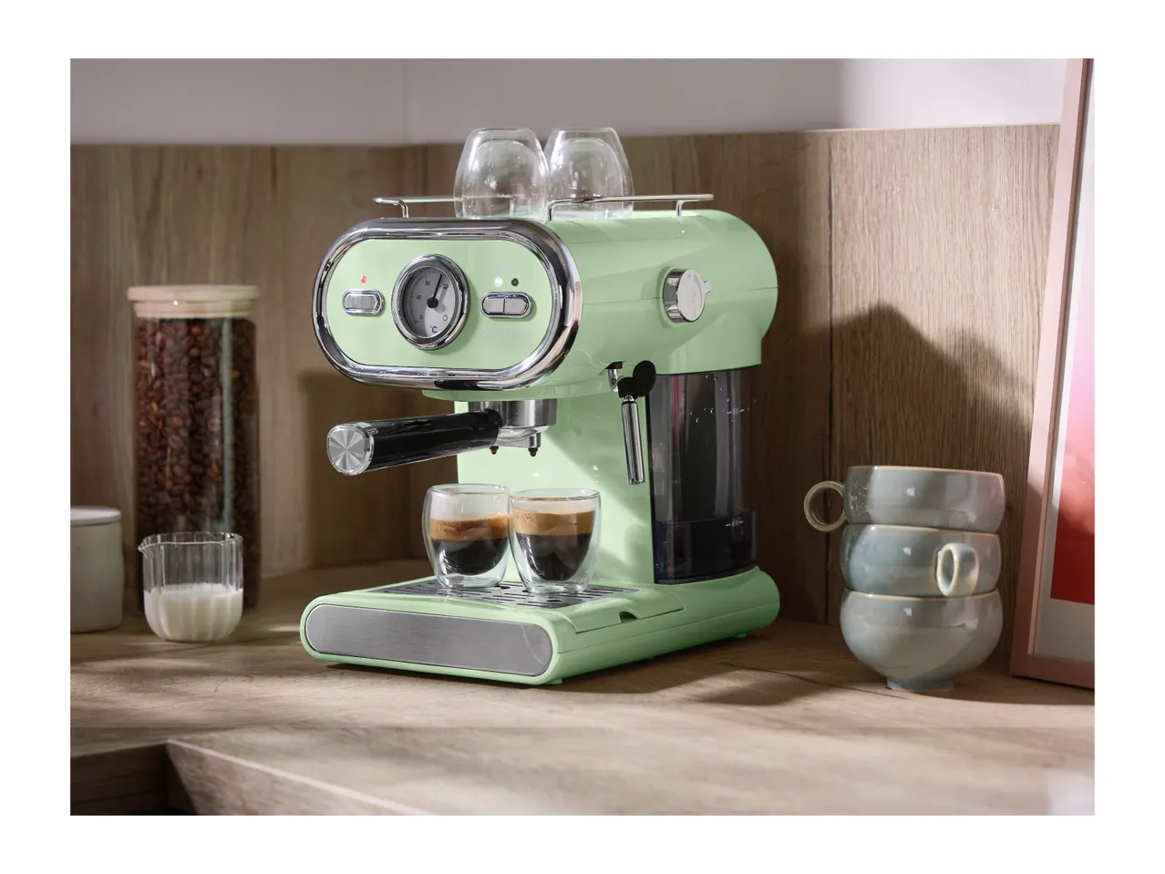 Light green espresso machine with two espresso cups, coffee beans, and milk on a wooden counter.