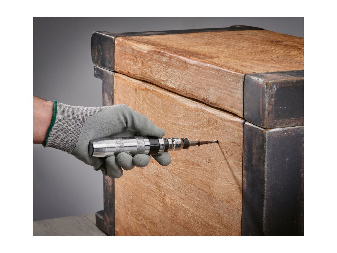 Gloved hand using an impact driver to remove a screw from a wooden chest.