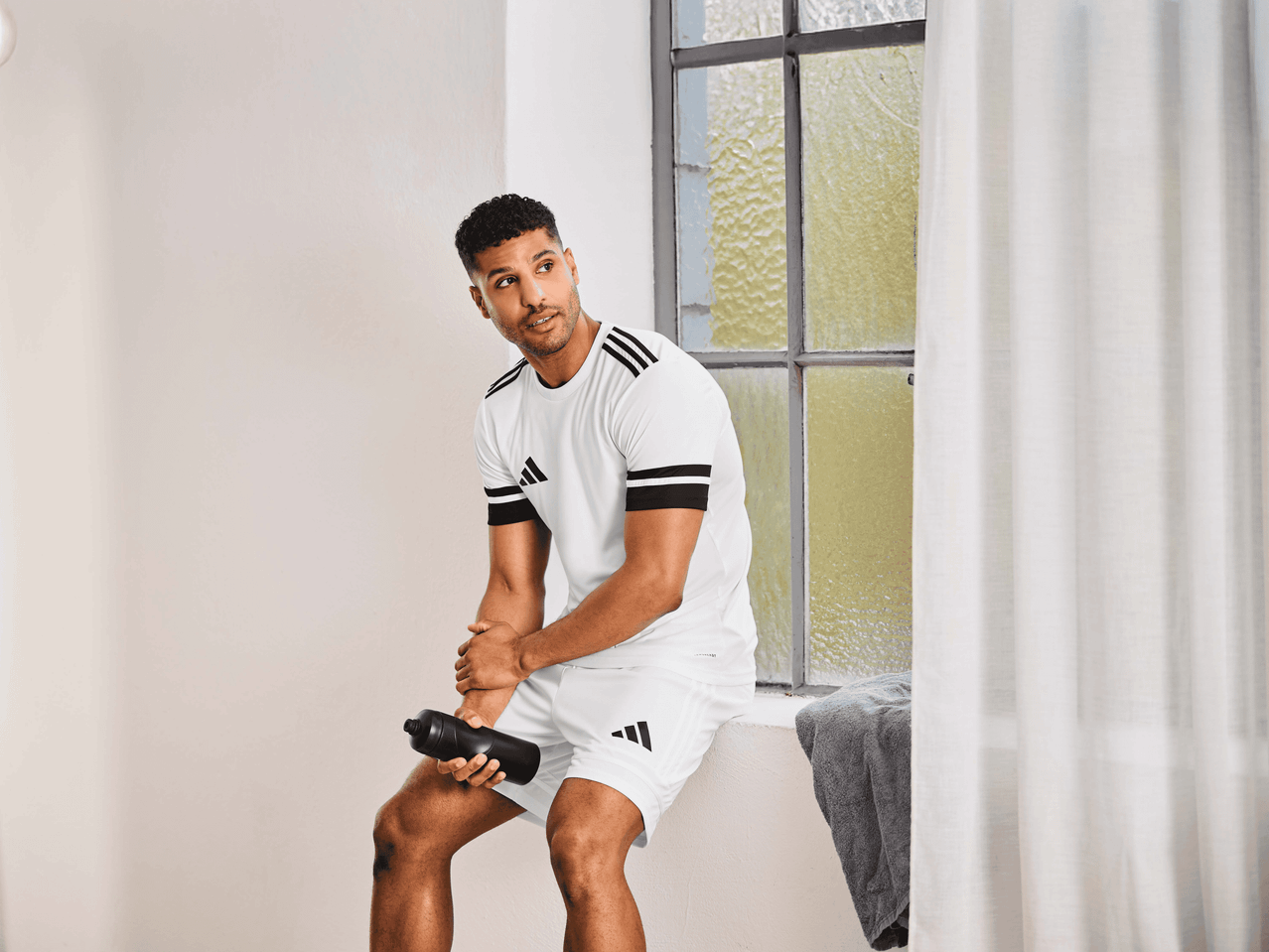 Mann in weißem Sport-T-Shirt und Shorts sitzt mit Wasserflasche am Fenster.