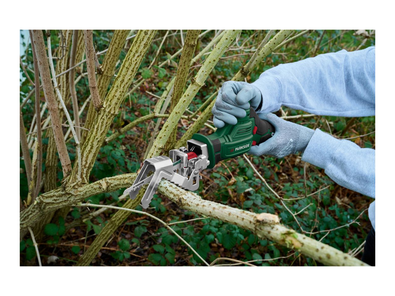 Person wearing gloves using a green cordless reciprocating saw to cut a tree branch.