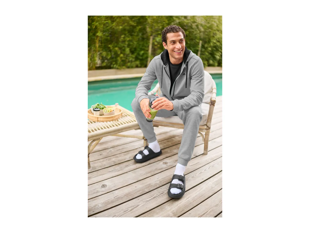 Man in grey tracksuit and black sandals with white socks, holding a green smoothie by a pool.