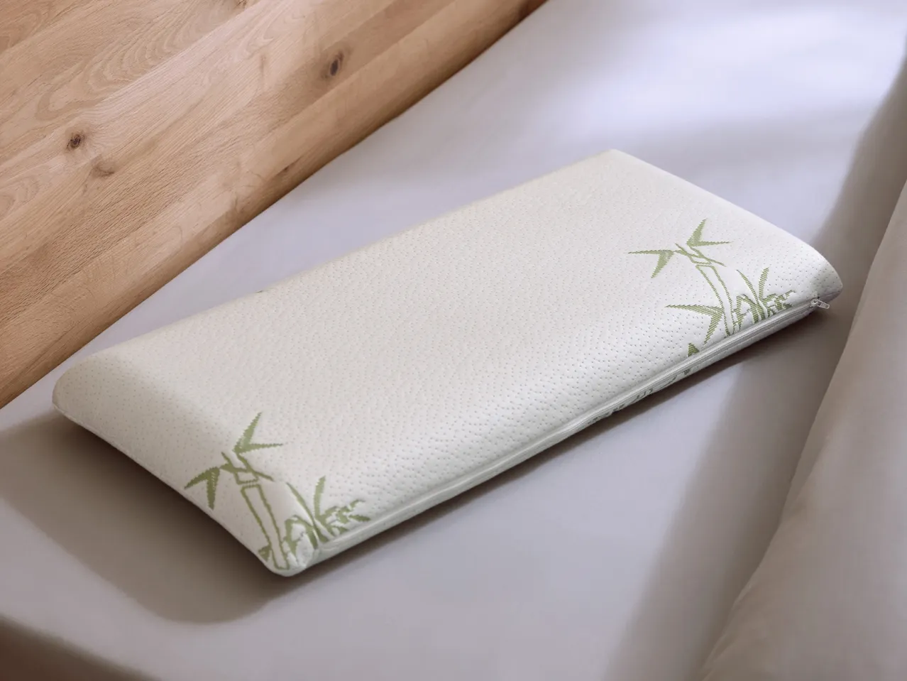 A white pillow with green bamboo patterns on a light grey sheet next to a wooden headboard.