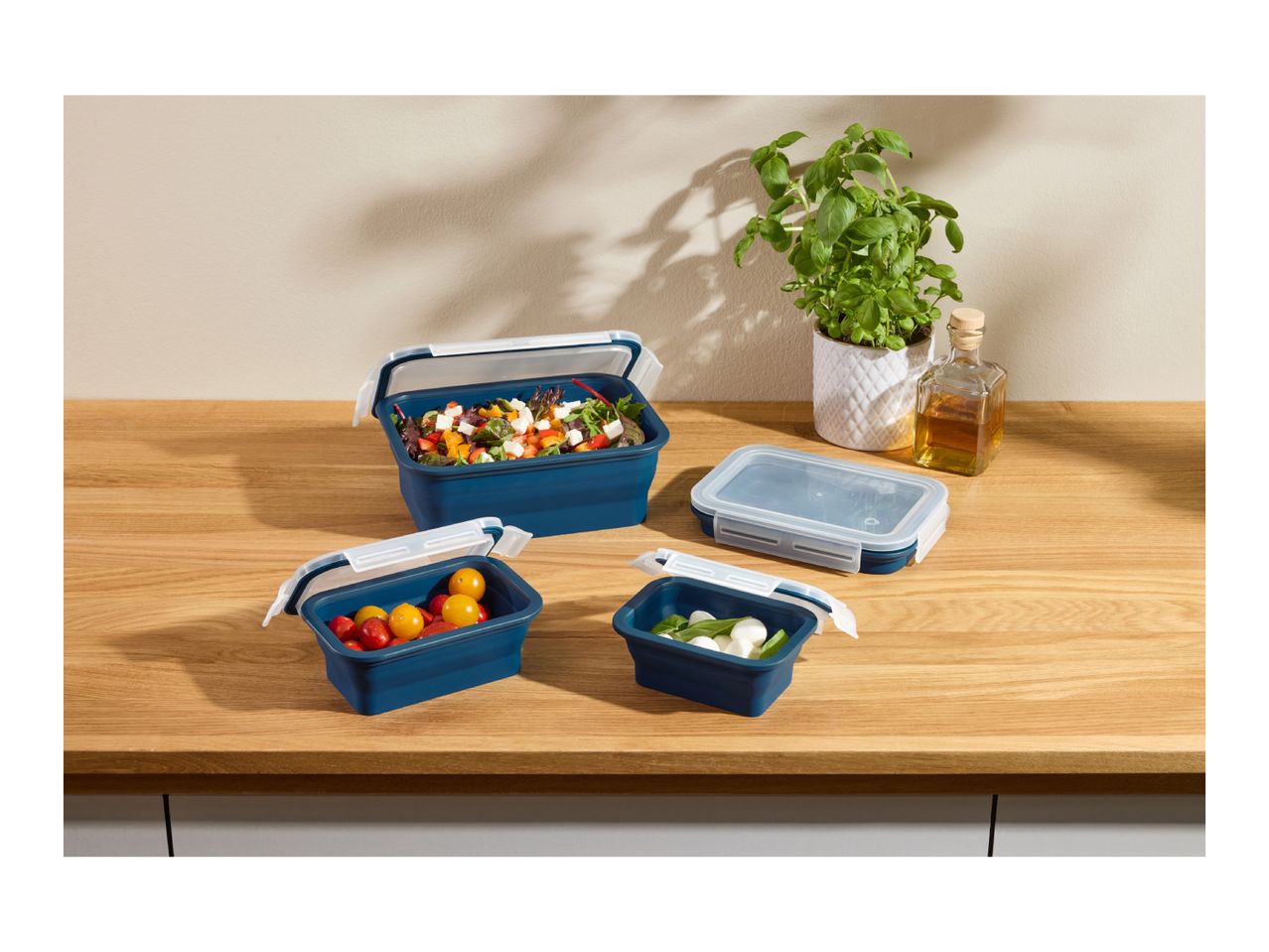Collapsible food containers with salad, tomatoes, and mozzarella on a wooden counter.