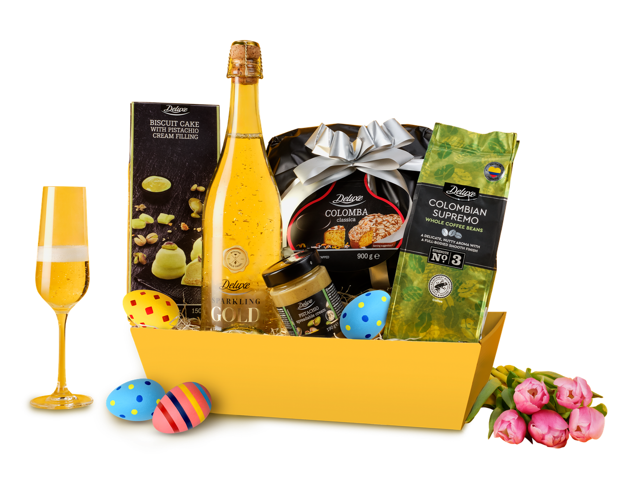 Un coș cadou de Paște cu o sticlă de vin spumant, prăjitură, cafea, cremă tartinabilă de fistic și ouă decorative.